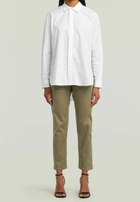 Chemise blanche à manches longues, légèrement ample. Un pantalon chino vert olive et des escarpins noirs à lanières complètent la tenue. Design simple, sans motifs.