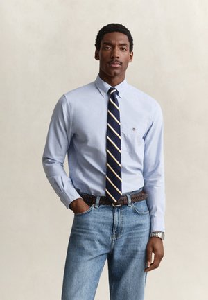 Homme portant une chemise bleu clair boutonnée, une cravate rayée bleu marine, un jean bleu, une ceinture marron tressée et une montre-bracelet argentée, debout avec une main dans la poche.