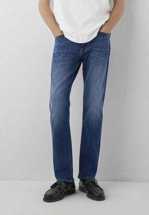 Uomo che indossa jeans blu a gamba dritta e scarpe di pelle nere, in piedi con le mani in tasca davanti a un muro semplice.