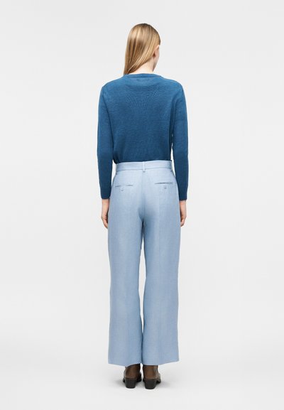 Γυναίκα με μακριά ξανθά μαλλιά που φοράει μπλε πουλόβερ και ανοιχτό μπλε παντελόνι ευρείας γραμμής, στέκεται με την πλάτη γυρισμένη σε λευκό φόντο.