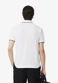 Polo bianco con accenti verdi nel colletto e nei polsini, caratterizzato da maniche corte e una vestibilità rilassata. Il tessuto sembra liscio e leggero.