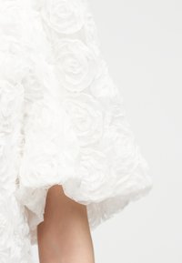 Λευκό ύφασμα με τρισδιάστατα λουλουδένια μοτίβα σε σχήμα τριαντάφυλλων, που καλύπτει μερικώς το χέρι ενός ατόμου πάνω σε απλό φωτεινό φόντο.