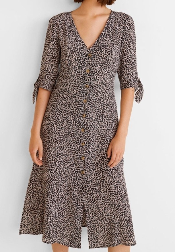 Femme portant une robe midi noire avec un petit imprimé floral blanc, col en V, boutons devant et manches trois-quarts nouées aux poignets.