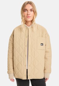 Beige quiltet jakke med knappelukking, ribbede mansjetter og brystlomme med et merket logo. Glatt tekstur med en rett passform.