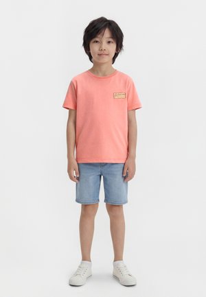 Enfant debout portant un t-shirt corail avec un patch "Summer at the Coast", un short en denim bleu clair, des baskets blanches, sur un fond blanc uni.