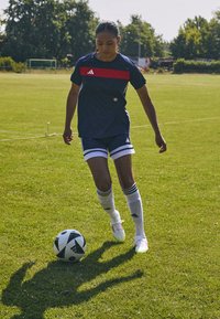 Adidas marinblå tröja med röd rand, vita shorts och knähöga vita strumpor, på en grön konstgräsplan. En svartvitt fotboll ligger i närheten.