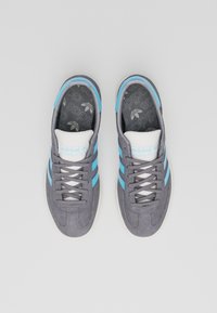 Grå mockasneakers med ljusblå ränder, vita accenter och en texturerad innersula med ett logomönster. Klassisk snörningsdesign.