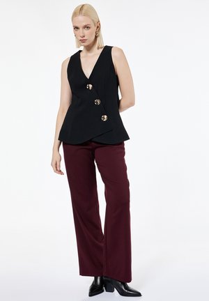 Kvinde iført sort ærmeløs top med store knapper, bordeaux vidde bukser og sorte ankellange støvler med hæl, stående mod hvid baggrund.