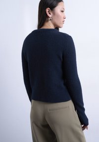 Γυναίκα που κοιτάζει πλάγια φορώντας ένα μπλε φορετό πουλόβερ και μπεζ παντελόνι, σε ένα απλό ανοιχτό φόντο.