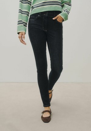 Person iført sorte skinny jeans, en grønstribet langærmet skjorte og brune Mary Jane-sko, stående med den ene hånd i lommen.
