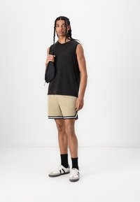 Chemise sans manches noire avec une poche avant, shorts en mesh beige avec des accents noirs, et des baskets grises et blanches ; debout avec un sac noir.