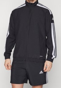 Black athletic jacket with white stripes, zip front, high collar, and fitted cuffs. Paired with matching black shorts and Adidas logo accents.