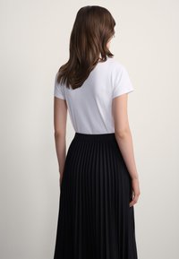 Kobieta o brązowych włosach, ubrana w biały krótki rękaw oraz wysoką plisowaną czarną spódnicę, stojąca tyłem na jednolitym tle.