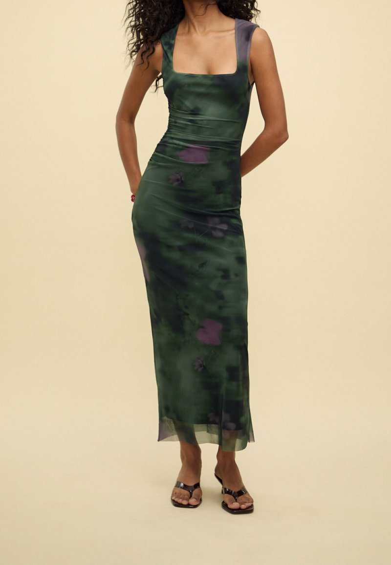 Femme portant une robe midi verte tie-dye sans manches et ajustée, et des sandales à talons hauts ouvertes noires, debout devant un fond beige.