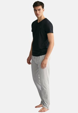 Junger Mann steht barfuß und trägt ein schwarzes kurzärmeliges Hemd und hellgraue lange Hosen vor einem schlichten weißen Hintergrund.