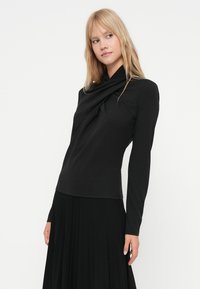 Sieviete ar blondiem matiem, tērpusies melnā ilgās piedurkņu blūzē ar drapētu kaklu un melnā plisētā svārkā, stāvot uz balta fona.