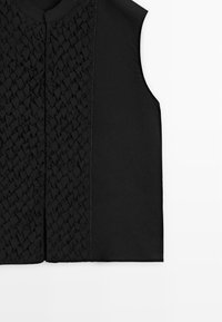 Černý bezrukávový oděv s propracovanou tkanou texturou na levé straně a hladkou látkou na pravé straně, zobrazený na bílém pozadí.