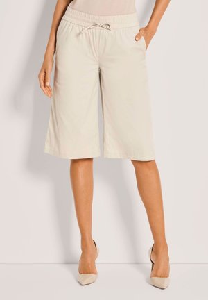 Frau trägt beige gekürzte, weit geschnittene Shorts mit Kordelzug an der Taille und passende beige High Heels, steht vor einem einfarbigen Hintergrund.