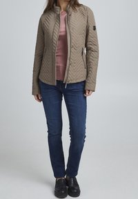 Beżowa pikowana kurtka w wzór jodełki, z kieszeniami na zamek i stojącym kołnierzem, noszona na różowej bluzce i niebieskich dżinsach. Czarne wsuwane buty.