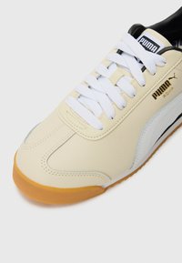Puma Roma sneakers in beige leather with white accents, featuring white laces, a textured finish, and a gum rubber sole. Gold logo detail.