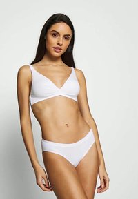 Γυναίκα με μακριά σκούρα μαλλιά στέκεται σε απλό φόντο φορώντας λευτό σουτιέν και αντίστοιχες λευκές εσώρουχες, ποζάροντας με το ένα χέρι χαλαρό.