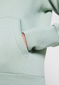 Felpa verde chiaro in tessuto texturizzato, con una grande tasca frontale e polsini a coste. Materiale morbido con una vestibilità comoda.