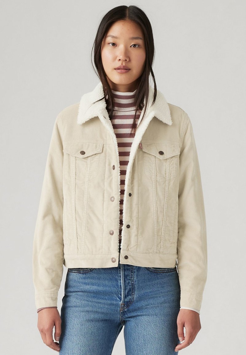Giacca di corduroy beige con collo in sherpa bianco, bottoni a scatto e due tasche sul petto. Indossata sopra una maglietta a righe a maniche lunghe.