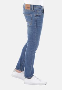 Pantalones vaqueros ajustados de mezclilla azul con una textura suave, que presentan un parche de cuero marrón en la parte trasera y bolsillos estándar, combinados con zapatillas blancas.