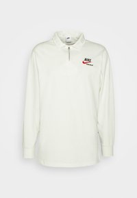 Polo de manga larga en color marfil, con cuello con cremallera, logo de Nike en rojo y negro, y una textura de tela suave y lisa.