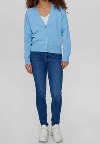 Ljusblå kofta i mjukt stickat material med v-ringning, knappfram, och hålmönster, kombinerad med figurnära jeans och vita sneakers.