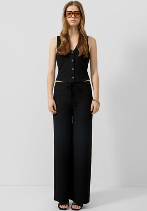 Femme aux longs cheveux bruns portant des lunettes de soleil ambrées, un haut noir sans manches à boutons, un pantalon large noir, et des chaussures noires ouvertes à l'avant.