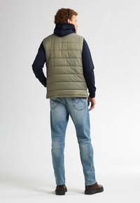 Olivengrüne Steppweste mit genähten Paneelen, kombiniert mit einem marineblauen Hoodie und hellblauen Jeans. Die Schuhe sind dunkelbraun mit einer dicken Sohle.