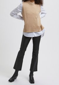 Beige mouwloze gebreide top over een wit overhemd met knopen, gecombineerd met zwarte flarebroeken en zwarte enkellaarsjes.