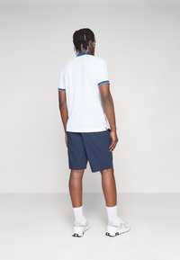 Chemise polo blanche avec des accents bleus, shorts navy et baskets légères. Le design est décontracté avec une coupe ample et des manches courtes.