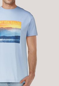 Camiseta de algodón azul claro con un estampado gráfico de montañas y agua, con acentos en amarillo y naranja, cuello redondo y mangas cortas.