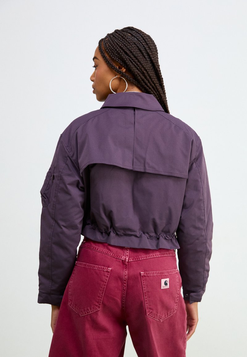 Chaqueta morada recortada con cuello estructurado y dobladillo elástico, combinada con pantalones burdeos de cintura alta con bolsillos traseros.