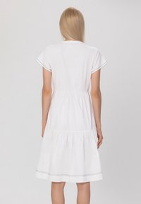 Robe en coton blanc avec des manches courtes, un ourlet à volants et des détails de couture contrastants. Texture lisse avec une coupe décontractée et un col arrondi.