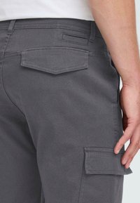 Close-up of gray cargo pants with flap pockets on the back and thigh, worn by a person with a white shirt and hand resting near the thigh pocket.