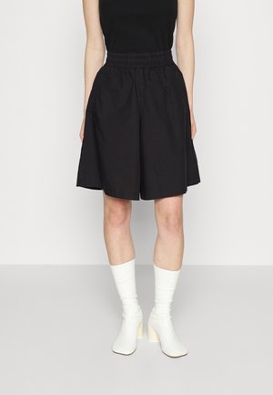 Kvinde iført sorte, højtaljede shorts til knæet med elastisk talje, kombineret med hvide hælede sokkestøvler og en sort ærmeløs top.