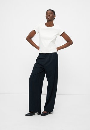 Femme debout, les mains sur les hanches, portant un haut blanc à manches courtes, un pantalon noir à jambes larges et des talons noirs sur un fond blanc.