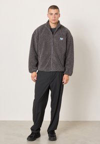 Giovane uomo in piedi, indossa una giacca in pile grigio scuro con cerniera, pantaloni neri larghi e sneakers nere su sfondo neutro.