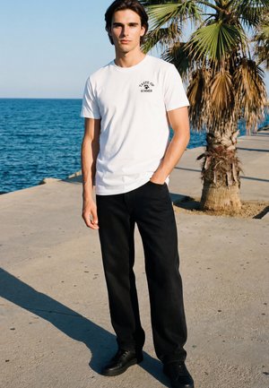 Junger Mann steht bei einer Palme am Meer und trägt ein weißes T-Shirt mit der Aufschrift "Taste of Summer", schwarze Hose und schwarze Schuhe.