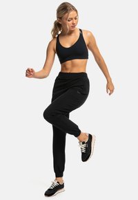 Sujetador deportivo negro combinado con pantalones jogger negros. Los joggers tienen un tobillo ajustado y bolsillos, complementados con zapatillas deportivas negras y rosas.