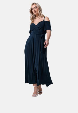 Blonde Frau trägt ein dunkelblaues schulterfreies Wickelkleid und silberne offene Absätze, steht in einer selbstbewussten Pose.