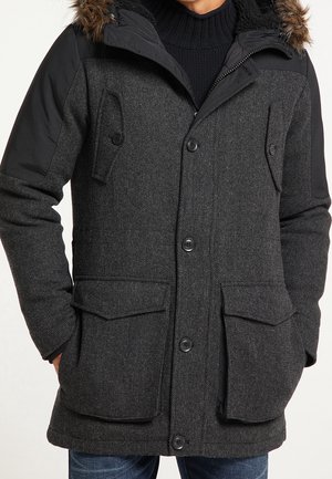 Manteau d'hiver texturé gris foncé avec des épaules en tissu noir, des poches à boutons et une capuche bordée de fourrure portée sur un pull noir.