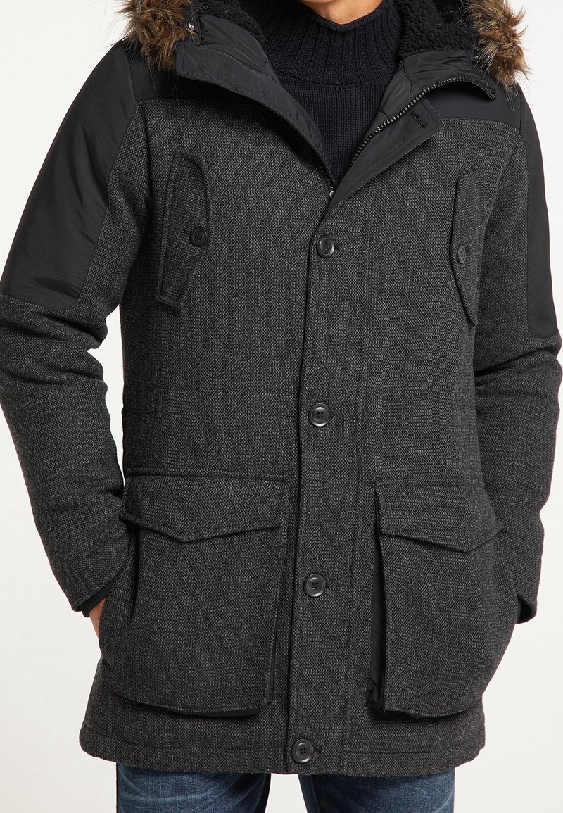 Manteau d'hiver texturé gris foncé avec des épaules en tissu noir, des poches à boutons et une capuche bordée de fourrure portée sur un pull noir.