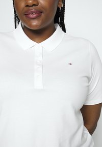 Femme à la peau foncée et aux cheveux tressés portant un polo blanc boutonné avec un petit logo sur la poitrine sur un fond uni.