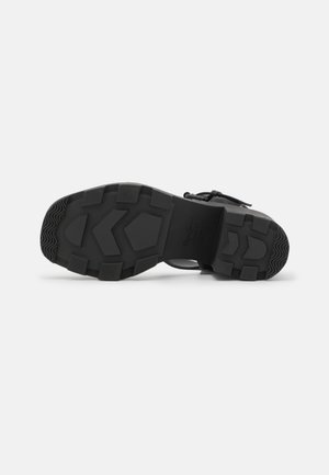 Semelle de botte noire avec des motifs de bande de roulement profonds et des grips en blocs, vue du dessous sur un fond blanc.