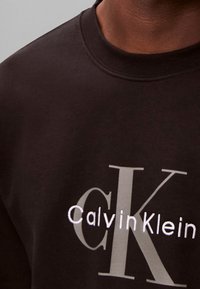 Chemise noire avec de grandes lettres grises "CK" et un logo brodé blanc "Calvin Klein" sur la poitrine, portée par une personne.