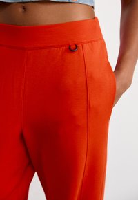 Close-up van een persoon in feloranje broek met een zwarte ringdetail bij de tailleband en een hand in de zak.